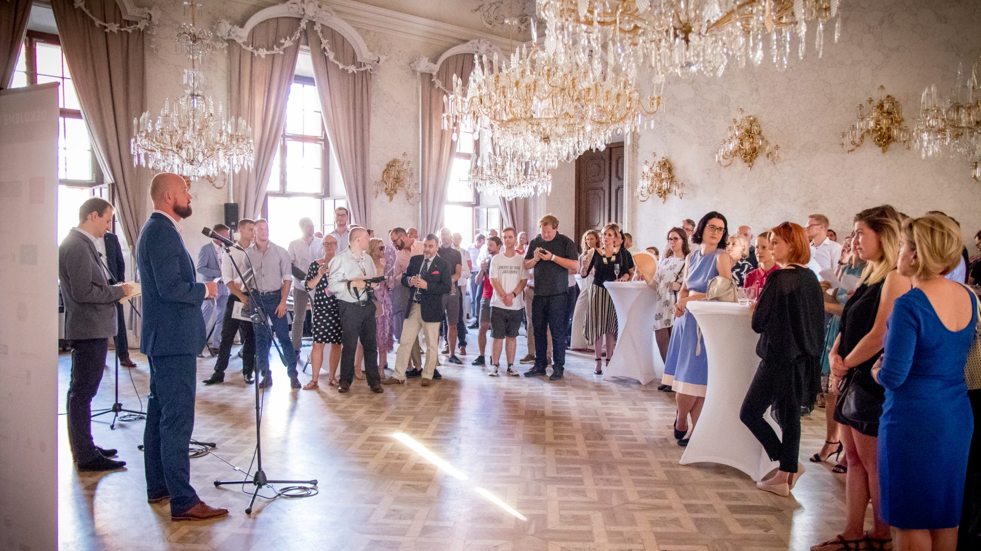 Photo of formal reception. People in formal clothes stanind in fancy room with chandeliers listening to a speech.