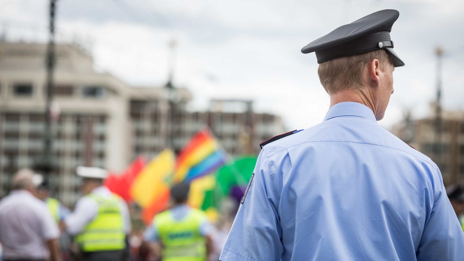 Fotografie policisty v uniformě, která se dívá do dálky. V pozadí jsou duhové vlajky a další policisté..